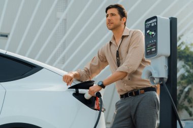 Genç adam, şehrin ticari otoparkındaki şarj istasyonundan elektrikli araba aküsünü şarj etmek için EV şarj aleti koymuş. Sürdürülebilir çevre dostu şehir yolculuğu için şarj edilebilir EV araba. Yararlı