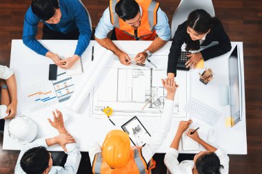 Mimari proje üzerinde birlikte çalışan, inşaat planını gözden geçiren ve toplantı masasında plan oluşturan çeşitli inşaat mühendisi ve müşteri gruplarından oluşan üst düzey bir pankart. Tedbirli