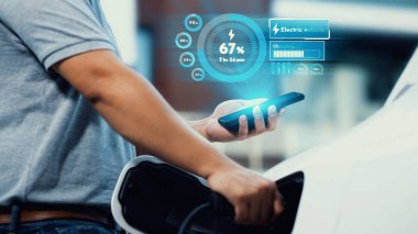 Modern yaşam tarzı adamı elektrikli arabasına EV şarj aleti yerleştiriyor ve akıllı telefondan EV batarya durum hologramını kontrol ediyor. Gelecekçi enerji sürdürülebilirliği ve elektrikli araba. İnceleyin