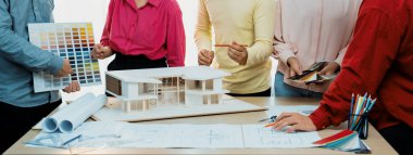 Profesyonel mimar takımının ev rengi seçimi hakkında beyin fırtınası yaparken, mimar da plan ve ev modelinin olduğu masadaki taslak belgeye yazı yazıyor. Değiştirilmiş.