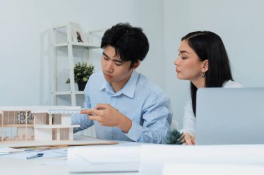 Profesyonel Asyalı erkek mimar ev modelinin uzunluğunu ölçmek için cetvel kullanırken, beyaz tenli genç meslektaşı ev modeliyle toplantı masasındaki verileri analiz etmek için dizüstü bilgisayar kullanıyor. Tertemiz..