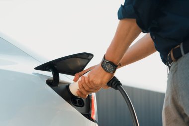 Genç adam, şehrin ticari otoparkındaki şarj istasyonundan elektrikli araba aküsünü şarj etmek için EV şarj aleti koymuş. Sürdürülebilir çevre dostu şehir yolculuğu için şarj edilebilir EV araba. Yararlı