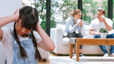 Stresli ve mutsuz genç kız köşeye kıvrılıp kulaklarını kapatarak arka planda ebeveyninin tartışmasını engelliyor. Evde aile içi şiddet ve travmatik çocukluk depresyona dönüşür. Eşzamanlama