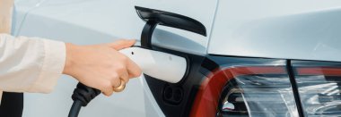 Genç bir kadın şarj aletini şarj istasyonundan elektrikli araba aküsünü şarj etmek için kullanmış. Sürdürülebilir çevre dostu seyahat kavramı için alternatif enerji ve şarj edilebilir EV araba. Panorama Uygulaması