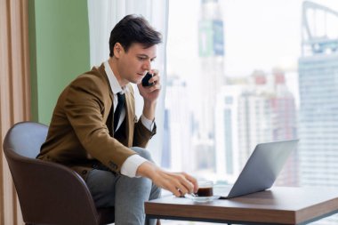 Telefon görüşmeleri sırasında dizüstü bilgisayarla çalışan profesyonel iş adamının portresi. Akıllı yönetici dizüstü bilgisayar ve kahve fincanı ile veri analizi yaparak arama yapıyor. Gökdelen manzaralı. Süslü.