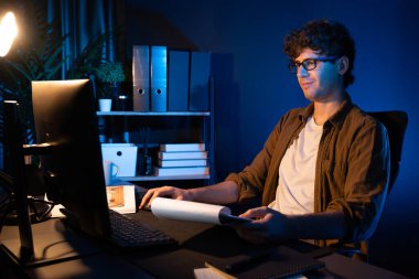 Genç işadamı, Neon Light Modern Ofis 'teki yeni proje surround çalışma masası için kağıt üzerinde tasarım düşünüyor. E-posta ile hediye göndermeden önce not defterine yeni bir iş düşünüyorum. Toplayıcı.