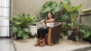 Modern genç kadın, tropikal bitkilerle çevrili, minimalist bir mimari beton stil yaz ekosistem bahçesinde uzaktan çalışıyor ya da dinleniyor. Blithe