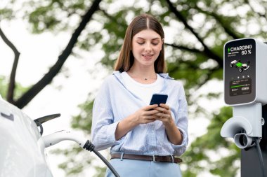 Ulusal Park veya Yaz Ormanı 'ndaki tatil gezisi sırasında EV şarj istasyonundan elektrik aküsü almak için akıllı telefon internet bankacılık uygulaması kullanan genç bir kadın. Yükselt