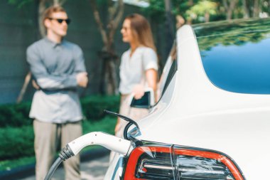 Genç çift yazın yeşil sürdürülebilir şehir bahçesinde EV elektrikli araba ile seyahat ederken, yeşil temiz yeniden şarj edilebilir elektrikli araç iç organları ile kentsel sürdürülebilirlik yaşam tarzını gösteriyor.