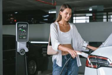 Genç bir kadın EV elektrikli arabasıyla alışveriş merkezinin otoparkına gidiyor şehir merkezinde elektrik iç organlarının yeşil temiz şarj edilebilir enerji ile sürdürülebilirlik yaşam tarzını gösteriyor.