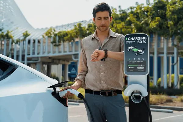 当电动汽车充电器充电时 年轻人在智能抽水机上查时间 以便从城市购物中心停车场的充电站充电 可再生能源电动汽车用于可持续的环境友好型城市旅行 — 图库照片