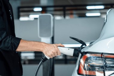 Genç adam EV elektrikli arabasıyla alışveriş merkezinin otoparkına gidiyor şehir merkezindeki şarj oluyor yeşil, temiz, yeniden şarj edilebilir elektrikli araç iç organları ile kentsel sürdürülebilirlik yaşam tarzını gösteriyor