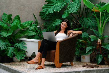 Modern genç kadın, tropikal bitkilerle çevrili, minimalist bir mimari beton stil yaz ekosistem bahçesinde uzaktan çalışıyor ya da dinleniyor. Blithe
