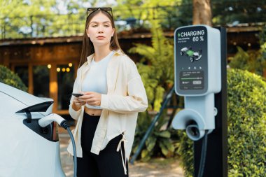 İlkbahar bahçesindeki açık kafede çalışan EV elektrikli araba şarjı, yeşil şehir sürdürülebilirliği ve çevre dostu EV otomobili için genç bir kadınla sürdürülebilir şehir seyahati elektrik ödüyor. Yararlı