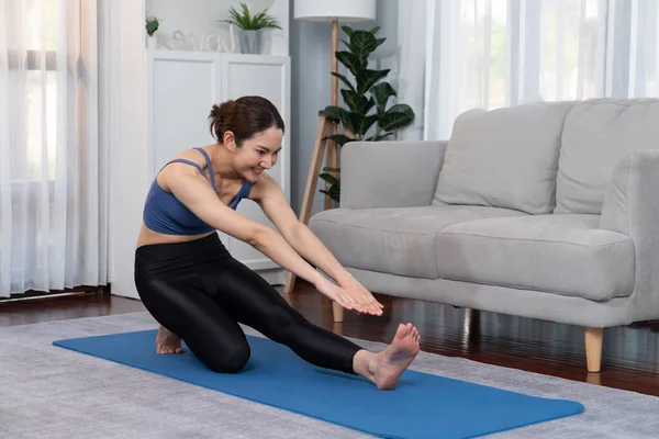 Asian woman in sportswear doing yoga exercise on fitness mat as her home workout training routine. Healthy body care and calm meditation in yoga lifestyle with comfortable and relaxation. Vigorous