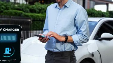 Modern iş adamı akıllı telefonun 3D hologramından batarya durumunu kontrol ederken EV arabasının şarj olan enerjisini kontrol ediyor. Gelecekteki EV otomotiv teknolojisinin yenilikçi teknolojik gelişimi. İnceleyin