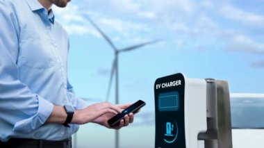 İş adamı akıllı telefondan batarya durum hologramını kontrol ederken rüzgar türbini jeneratöründen gelen alternatif temiz enerjiyi kullanarak çevre dostu EV arabasını şarj ediyor. İnceleyin
