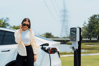 EV araba aküsünü şarj eden genç bir kadın elektrik şebekesine bağlı elektrik santralinde çevre dostu araç kullanımı için elektrik endüstrisi olarak telefonla konuşurken. Yararlı