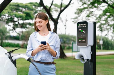 Genç kadın EV şarj istasyonundan EV elektrikli araç aküsünü şarj ediyor ve şehir parkındaki elektriği ödemek için akıllı telefon internet bankacılığını kullanıyor. EV arabalı çevre dostu bir araç. Yükselt