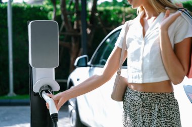 Genç bir kadın yazın yeşil sürdürülebilir şehir bahçesinde EV elektrikli arabasıyla seyahat ediyor. Kentsel sürdürülebilirlik yaşam tarzı yeşil temiz yeniden şarj edilebilir enerji elektrikli BEV araç iç organları
