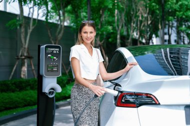 Genç bir kadın yazın yeşil sürdürülebilir şehir bahçesinde EV elektrikli arabasıyla seyahat ediyor. Kentsel sürdürülebilirlik yaşam tarzı yeşil temiz yeniden şarj edilebilir enerji elektrikli BEV araç iç organları