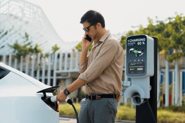 Genç adam, şehrin ticari otoparkındaki şarj istasyonundan elektrikli araba aküsünü şarj etmek için EV şarj aleti koymuş. Sürdürülebilir çevre dostu şehir yolculuğu için şarj edilebilir EV araba. Yararlı