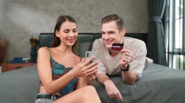 Genç çift, kredi kartıyla ödemek için internet üzerinden ödeme uygulaması ve dijital cüzdan kullanarak yatak odasında oturuyor. Mobil internet üzerinden alışveriş ve modern alımlar. Düzenle
