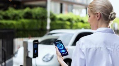 Akıllı telefon akü durumu arayüzünü elinde tutan iş kadını akıllı EV mobil uygulama ile otomobil parkındaki şarj istasyonundan elektrik şarj eden çevre dostu EV araba. İnceleyin