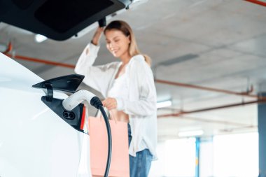 Genç bir kadın EV elektrikli arabasıyla alışveriş merkezinin otoparkına gidiyor şehir merkezinde elektrik iç organlarının yeşil temiz şarj edilebilir enerji ile sürdürülebilirlik yaşam tarzını gösteriyor.