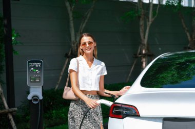 Genç bir kadın yazın yeşil sürdürülebilir şehir bahçesinde EV elektrikli arabasıyla seyahat ediyor. Kentsel sürdürülebilirlik yaşam tarzı yeşil temiz yeniden şarj edilebilir enerji elektrikli BEV araç iç organları