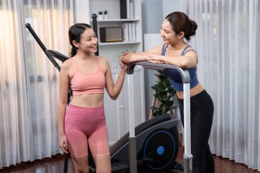 Enerjik ve güçlü atletik Asyalı kadın evde spor arkadaşı ya da antrenörü ile birlikte eliptik koşu makinesiyle koşuyor. Formda fiziğin takibi ve sağlıklı yaşam tarzına bağlılık. Kuvvetli