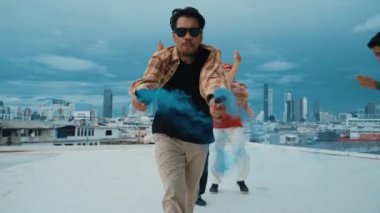 Bir grup çok kültürlü hippi çatıda işaret fişeği çalıyor. Zenci dumanı tutan çekici bir sokak dansçısı hip-hop müzik eşliğinde break dans yapıyor. Açık hava sporu 2024. Sokak dansı. hiphop.