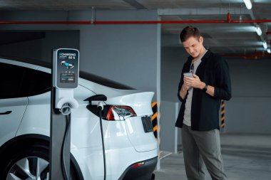 Genç adam EV elektrikli arabasıyla alışveriş merkezinin otoparkına gidiyor şehir merkezindeki şarj oluyor yeşil, temiz, yeniden şarj edilebilir elektrikli araç iç organları ile kentsel sürdürülebilirlik yaşam tarzını gösteriyor