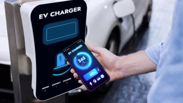 スマートフォンを持つビジネスマンはスマートEvモバイルアプリケーションによるバッテリーステータスインターフェースを表示し エコフレンドリーなEv車は駐車場の充電ステーションから電気を充電します Peruse — ストック動画