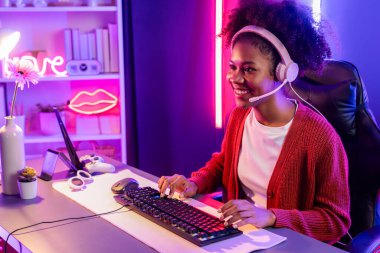 Oyun flama, Afrikalı kız Esport yetenekli takımıyla online kavga ediyor. Neon renkli aydınlatma odasında kulaklık takıyor. Rakiplerini kazanmak için strateji planlayan diğer oyuncularla konuşmak. Tatlandırıcı.