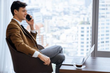 Gökdelen manzaralı pencerenin yanında otururken yakışıklı iş adamının müdürle telefon görüşmesi yaptığı yakın plan. Yönetici, telefon ve dizüstü bilgisayarla konuşuyor. Önüne bak. Süslü.