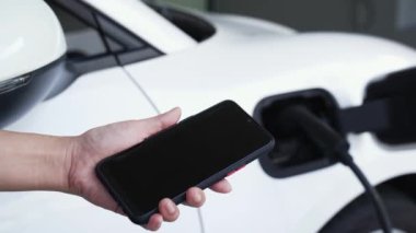 Akıllı telefon ekranı akıllı EV mobil uygulama üzerinde pil durumu EV araba garajdaki ev şarj istasyonu ile elektrik şarj ederken. Geleceğin yenilikçi EV arabası ve enerji sürdürülebilirliği. İnceleyin