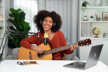 Genç Afrikalı Amerikalı müzisyenin sunucu kanalı gitar çalıyor, şarkı söylüyor, stüdyoda dizüstü bilgisayardan yayın yapıyor. Kulaklık dekorasyonu ve mikrofon kaydı. Tatlandırıcı.