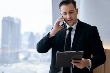 Şirketin yetenekli iş adamı telefonla konuşuyor ve pencerenin yanında dururken verilere bakıyor. Gökdelen manzarası gösteriyor. Telefonla çalışan ciddi meşgul bir yönetici. Modern işyeri, ofis. İş için. Süslü.