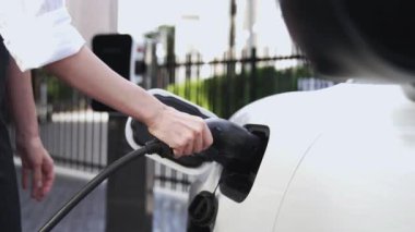 İş kadını akıllı EV şarj aleti takıyor ve şehir otoparkındaki EV şarj istasyonundan elektrikli araba şarj ediyor. Geleceksel sürdürülebilir güç ve EV araba kullanımı. İnceleyin