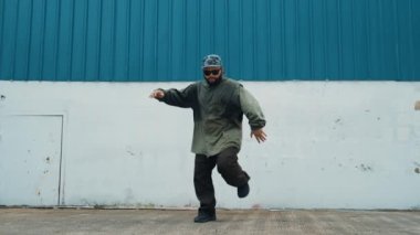 Sokak ortasında mavi duvarlı hippi dansçı ayak basamağı. Asyalı break dansçı kollarını açarken sokak dansı yapıyor. Hip hop koreografı enerjik bir dans sergiliyor. Açık hava sporu 2024 hiphop..