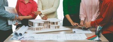 Profesyonel mimar takımının ev rengi seçimi hakkında beyin fırtınası yaparken, mimar da plan ve ev modelinin olduğu masadaki taslak belgeye yazı yazıyor. Değiştirilmiş.