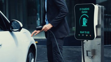 İş adamı elektrikli arabasını şehir merkezindeki şarj istasyonundan ya da otoparktan şarj ediyor. Şehir yaşam tarzında alternatif temiz enerji kullanan çevre dostu şarj edilebilir araba. İnceleyin.
