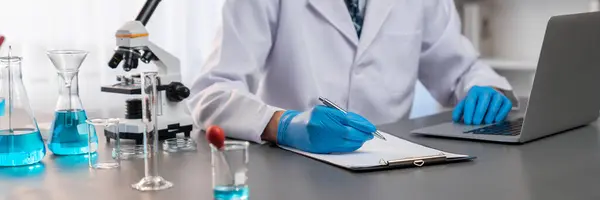 Closeup hand recording clinical data result after conducting chemical experiment in medical laboratory for vaccine drug or antibiotic. Scientific chemistry lab and medicine research concept. Neoteric