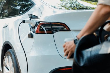 Genç adam baharda açık yeşil şehir parkındaki şarj istasyonundan elektrikli araba aküsünü şarj ediyor. Sürdürülebilir çevre dostu kentsel seyahat tarzı için şarj edilebilir EV araba. Yararlı