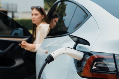 Genç bir kadın EV otomobil şarj istasyonu Green City Park 'ta elektrik ödemek için akıllı telefonu kullanıyor. Modern çevre ve EV araçlı sürdürülebilir şehir yaşam tarzı. Yararlı