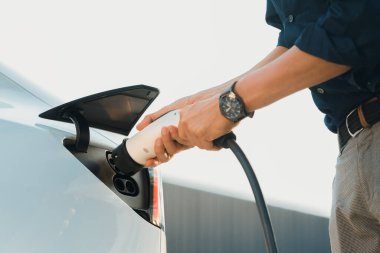 Genç adam, şehrin ticari otoparkındaki şarj istasyonundan elektrikli araba aküsünü şarj etmek için EV şarj aleti koymuş. Sürdürülebilir çevre dostu şehir yolculuğu için şarj edilebilir EV araba. Yararlı