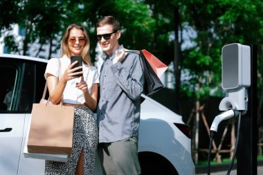 Genç çift yazın yeşil sürdürülebilir şehir bahçesinde EV elektrikli araba ile seyahat ederken, yeşil temiz yeniden şarj edilebilir elektrikli araç iç organları ile kentsel sürdürülebilirlik yaşam tarzını gösteriyor.