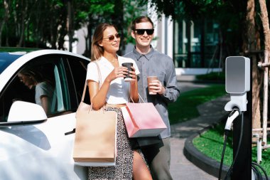 Genç çift yazın yeşil sürdürülebilir şehir bahçesinde EV elektrikli araba ile seyahat ederken, yeşil temiz yeniden şarj edilebilir elektrikli araç iç organları ile kentsel sürdürülebilirlik yaşam tarzını gösteriyor.