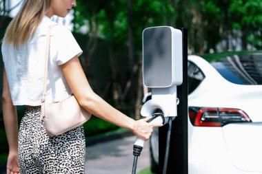 Genç bir kadın yazın yeşil sürdürülebilir şehir bahçesinde EV elektrikli arabasıyla seyahat ediyor. Kentsel sürdürülebilirlik yaşam tarzı yeşil temiz yeniden şarj edilebilir enerji elektrikli BEV araç iç organları